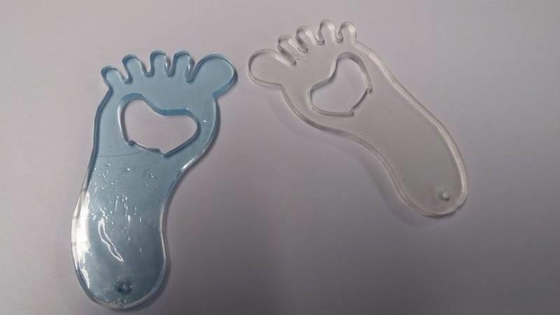 Dwa otwieracze do butelek w kształcie stóp, jeden niebieski i jeden przezroczysty, z wyciętym sercem w środku, leżące na białym tle.