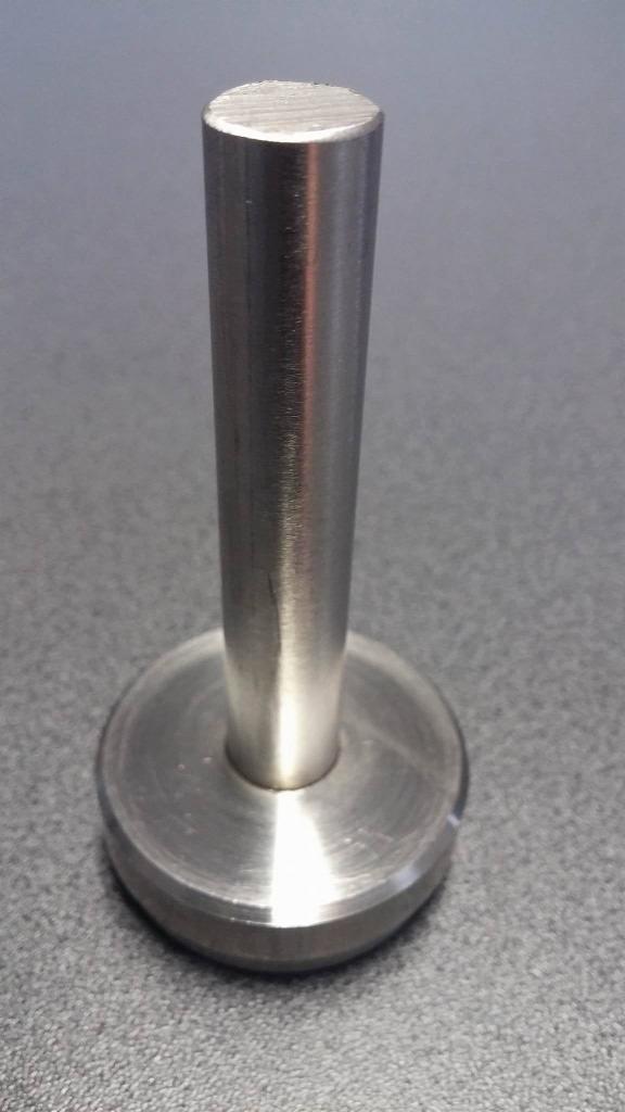 Metalowy trzpień z okrągłą podstawą, widoczne ślady obróbki mechanicznej na powierzchni, leżący na szarym blacie.