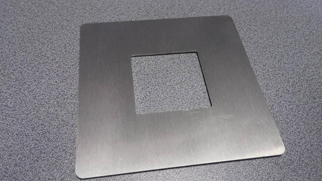 Kwadratowa, metalowa płyta z wyciętym kwadratowym otworem pośrodku, leżąca na szarej, teksturowanej powierzchni.