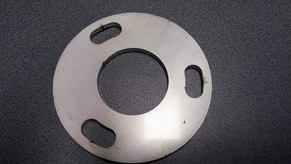 Szlifowany, metalowy dysk z trzema podłużnymi otworami i centralnym okrągłym otworem, leżący na ciemnoszarym, teksturowanym podłożu.