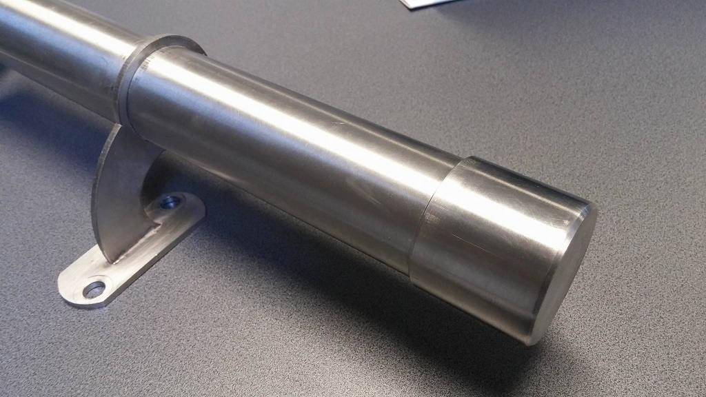 Szczegółowe ujęcie polerowanej, stalowej rury z zamocowanym wspornikiem, leżącej na szarej powierzchni.