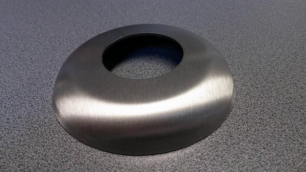 Szczotkowana, metalowa rozeta o okrągłym kształcie z otworem centralnym, leżąca na szarej, teksturowanej powierzchni.