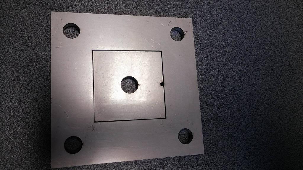 Szara, metalowa płyta z czterema otworami w rogach i wyciętym kwadratem wewnątrz, z centralnym otworem i małym otworem w rogu kwadratu, leżąca na ciemnoszarym, teksturowanym tle.