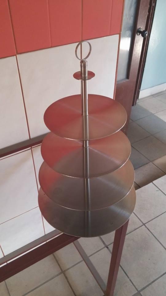 Metalowa, czteropoziomowa etażerka z okrągłymi półkami, stojąca na metalowym stelażu, widoczna na tle kafelków i drzwi.