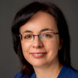 Portret uśmiechniętej kobiety w okularach z fioletowymi oprawkami, ubrana w niebieską bluzkę, na jednolitym szarym tle.