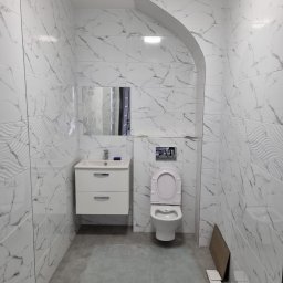 PHU Edyta Golec - Nowoczesna łazienka z białymi marmurkowymi płytkami ściennymi, szarą podłogą, białym zlewem z szafką, lustrem i toaletą z otwartą klapą; widoczne drobne niedociągnięcia na podłodze i przy toalecie.