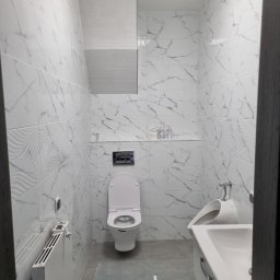 PHU Edyta Golec - Wykończona łazienka z białymi płytkami imitującymi marmur, wisząca toaleta z otwartą klapą, umywalka z szafką i otwartą szufladą oraz grzejnik elektryczny z wystającym kablem.