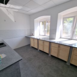 PHU Edyta Golec - Wnętrze kuchni w trakcie wykańczania: szare płytki podłogowe, zabudowa kuchenna z drewnianymi frontami żaluzjowymi pod oknami z zaokrąglonymi wnękami, białe ściany z białymi płytkami nad blatem.