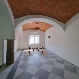 PHU Edyta Golec - Wnętrze odnowionego pomieszczenia z ceglanym sklepieniem łukowym, szachownicą z szarych płytek na podłodze i kamienną kolumną. Dwie ławki stoją pod oknem, a na podłodze leżą kartony zabezpieczające.