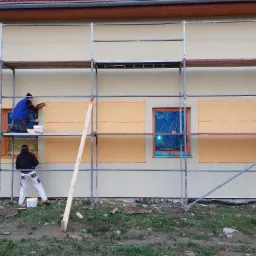 Prace na elewacji budynku z użyciem rusztowania; robotnicy aplikują warstwę tynku na fragmenty ściany wokół okien zabezpieczonych folią.