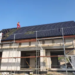 Dwóch pracowników na rusztowaniu montuje ciemną dachówkę na nowym domu z pustaków, widoczny fragment więźby dachowej.