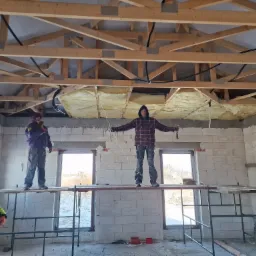 Trzech mężczyzn na rusztowaniach w surowym wnętrzu budynku z pustaków, montujących izolację poddasza pod drewnianą więźbą dachową