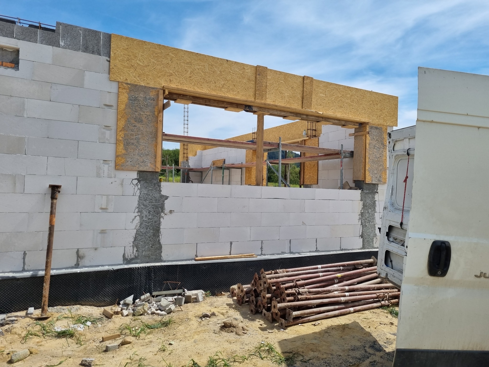 Wznoszenie ściany z bloczków betonowych z otworem okiennym wzmocnionym drewnianym szalunkiem, widoczne podpory budowlane i fragment samochodu dostawczego.
