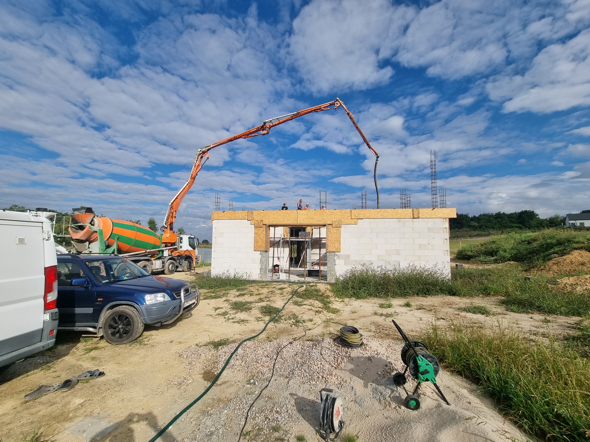 Wylewka betonu na strop budynku z bloczków, widoczna pompa do betonu, betoniarka i pracownicy na górze, w tle błękitne niebo z chmurami.