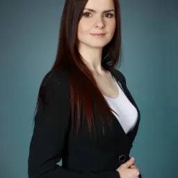 Portret kobiety z długimi, rudymi włosami, ubranej w czarną marynarkę i białą bluzkę, pozującej na tle jednolitego, błękitnego tła.