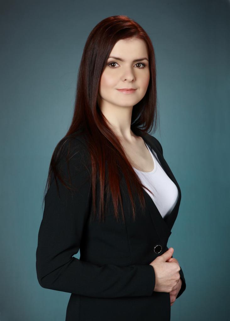 Portret kobiety z długimi, rudymi włosami, ubranej w czarną marynarkę i białą bluzkę, pozującej na tle jednolitego, błękitnego tła.