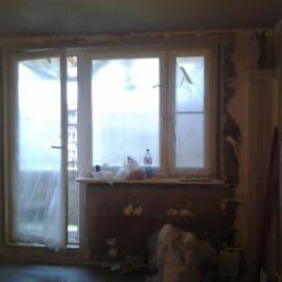 Renomex - Pokój w trakcie prac przygotowawczych przed wykończeniem: okna zakryte folią, ściany w surowym tynku, widoczne narzędzia i materiały budowlane, wnętrze w stanie przed malowaniem lub tapetowaniem.