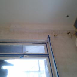 Renomex - Przygotowanie do malowania: okno zabezpieczone niebieską taśmą malarską, ściana po szpachlowaniu z widoczną fakturą, puszka farby w tle.