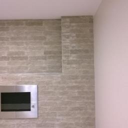 Renomex - Ściana wykończona panelami imitującymi kamień, z wbudowanym metalowym wkładem kominkowym, widok na fragment kuchni w tle.