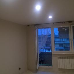 Renomex - Jasne wnętrze po remoncie z widokiem na balkon i budynek mieszkalny, świeżo pomalowane ściany w odcieniach beżu, biały grzejnik pod oknem, zamontowane karnisze i oświetlenie sufitowe.