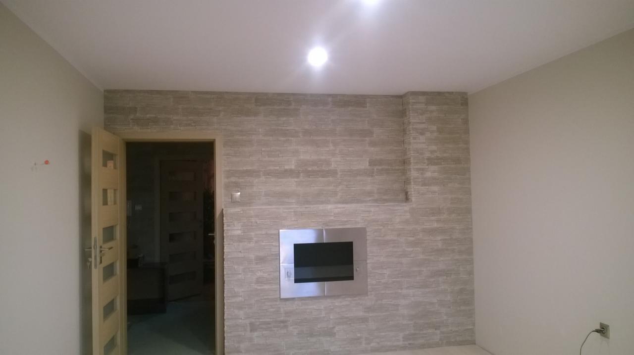 Ściana wykończona szarą okładziną imitującą kamień z wbudowanym biokominkiem w metalowej obudowie, widoczny fragment otwartych drzwi z przeszkleniami.