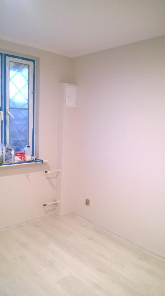 Wnętrze pokoju po odświeżeniu ścian na jasny odcień, okno zabezpieczone taśmą malarską, podłoga wyłożona panelami w odcieniach bieli.