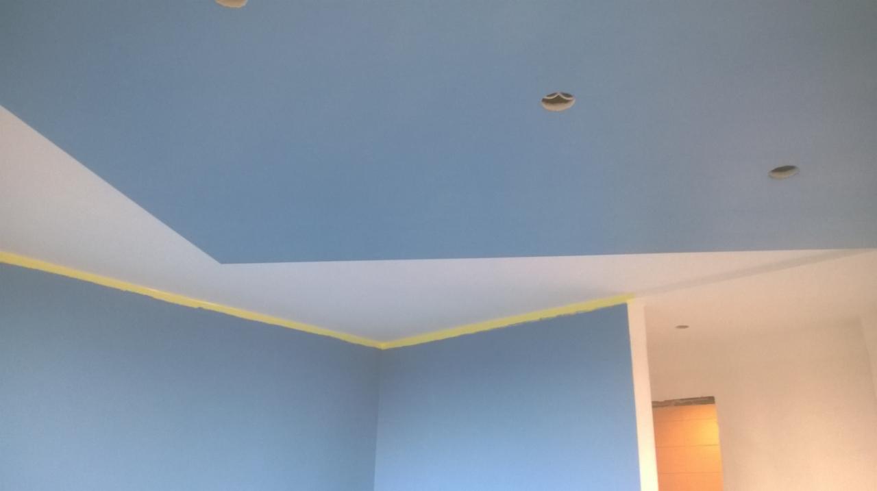 Pomieszczenie po malowaniu ścian i sufitu na kolor niebieski, z żółtą taśmą malarską zabezpieczającą krawędzie, widoczne otwory na oświetlenie w suficie.