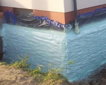 Niska partia ściany budynku zabezpieczona grubą warstwą jasnoniebieskiego materiału izolacyjnego, wystający fragment rury aplikującej, widoczne trawy w dolnej części kadru.