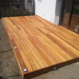 Nowoczesny taras z drewna egzotycznego, z wbudowanymi lampkami LED, przylegający do budynku z drzwiami tarasowymi.