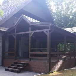 Drewniana altana przylegająca do kamiennego budynku z dachem krytym gontem, otoczona zielenią drzew.