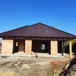 Niedokończony budynek z cegły z brązowym dachem z dachówki ceramicznej w słoneczny dzień. Widoczne krokwie i fragment komina. Teren wokół budynku w stanie surowym.