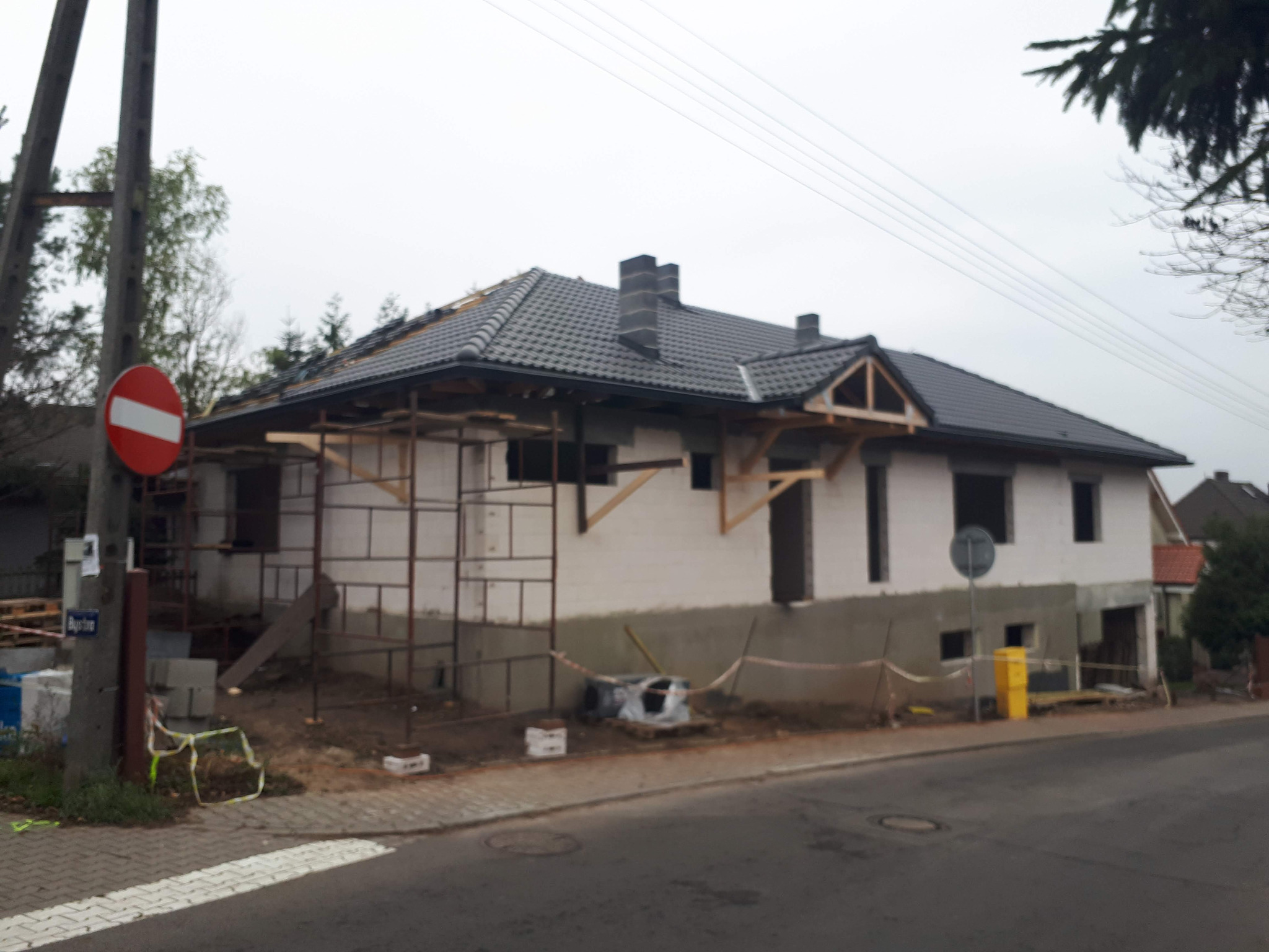 Dom w trakcie budowy z częściowo położonym ciemnoszarym dachem, widoczne rusztowania i niezakończone elewacje, znak drogowy zakazu w tle.