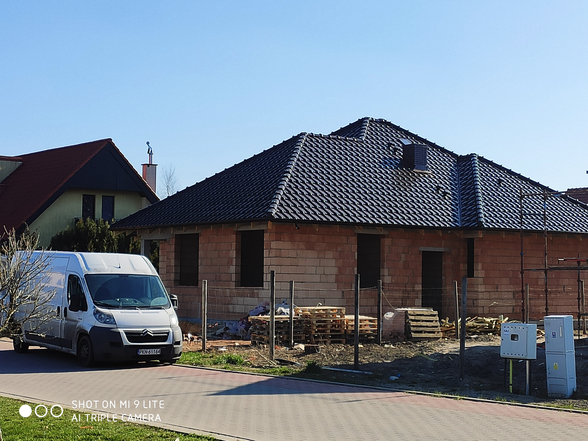 Dom jednorodzinny w trakcie budowy, z nowym, ciemnym dachem z dachówki ceramicznej, widoczne rusztowania, palety z materiałami budowlanymi i zaparkowany biały samochód dostawczy.