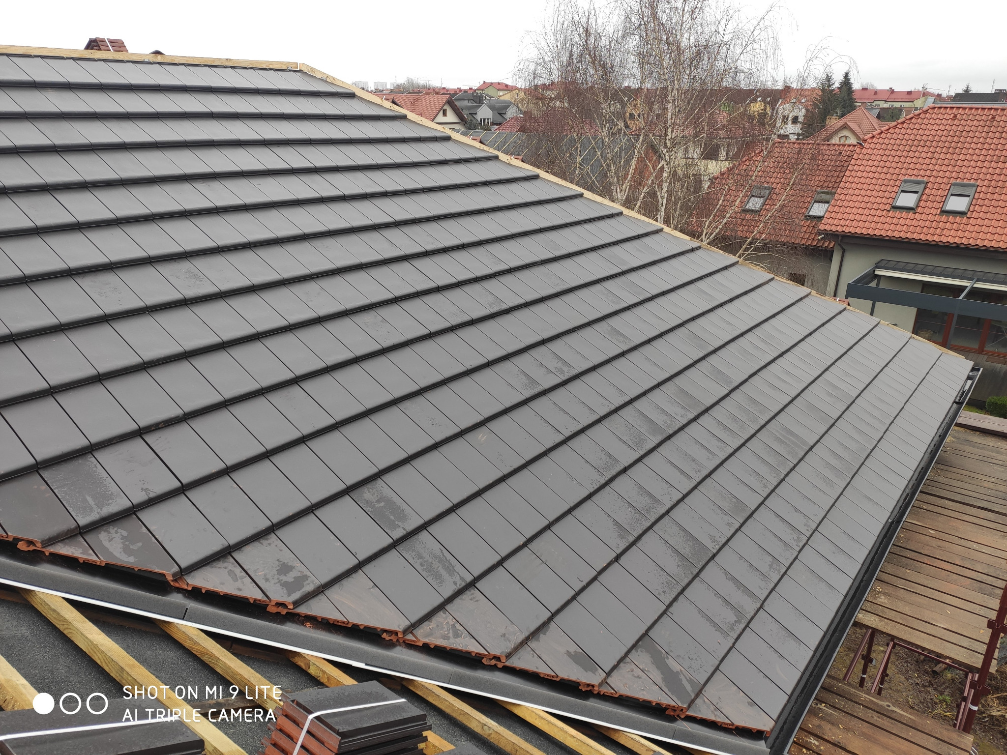 Nowo położona, grafitowa dachówka ceramiczna na konstrukcji drewnianej, widoczne elementy więźby, w tle zabudowa jednorodzinna.