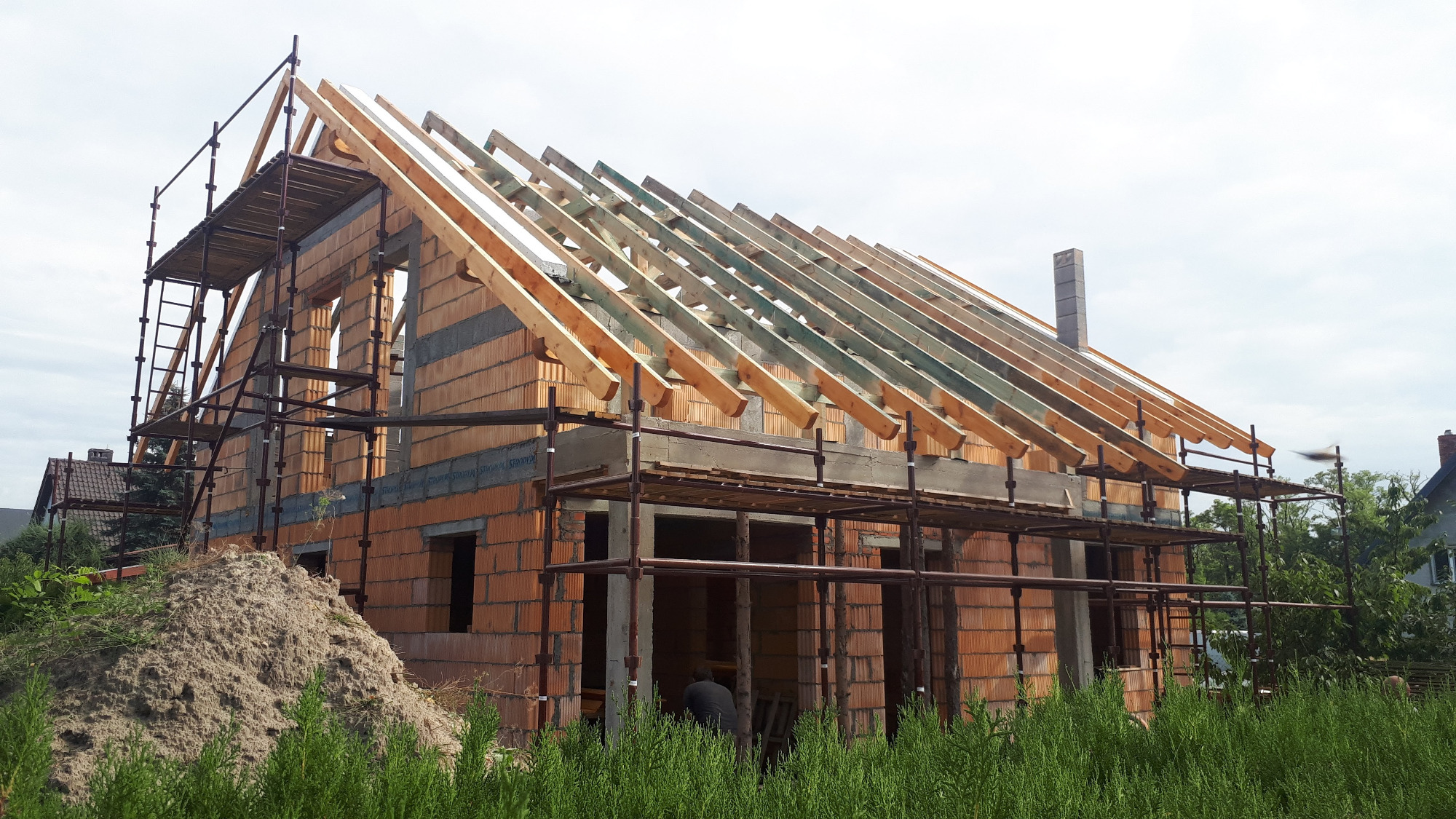 Budowa domu jednorodzinnego w trakcie montażu drewnianej konstrukcji dachu, widoczne rusztowania i stos cegieł.