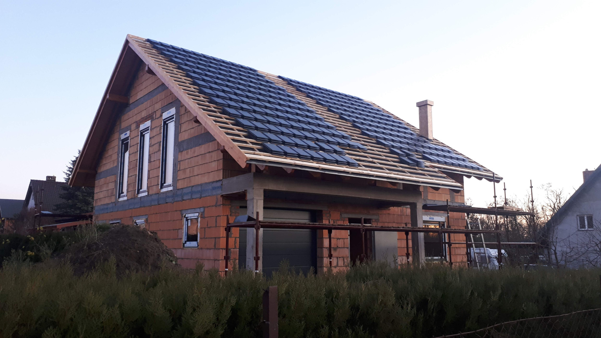 Dom jednorodzinny w trakcie budowy, widoczny fragment dachu z ułożoną częścią czarnej dachówki ceramicznej, niezakończona konstrukcja garażu, ściany z czerwonej cegły dziurawki.