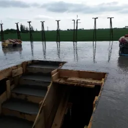 Zalany betonem fundament z drewnianym szalunkiem schodów, widoczne wystające pręty zbrojeniowe i plandeki z cegłami w tle, pochmurne niebo.