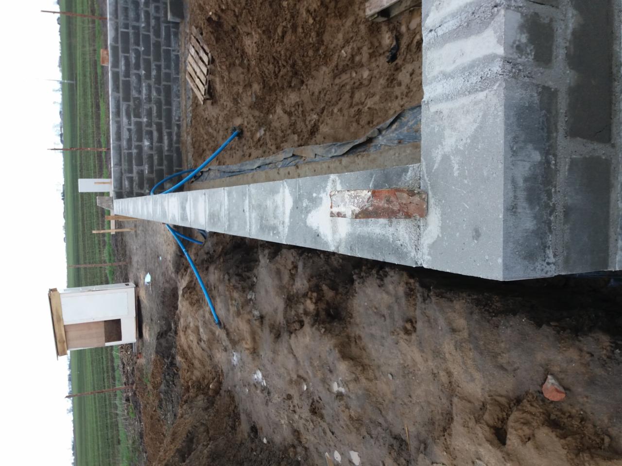 Wylewka fundamentowa z wmurowaną czerwoną cegłą, widoczne niebieskie rurki i ściana z bloczków betonowych w tle.