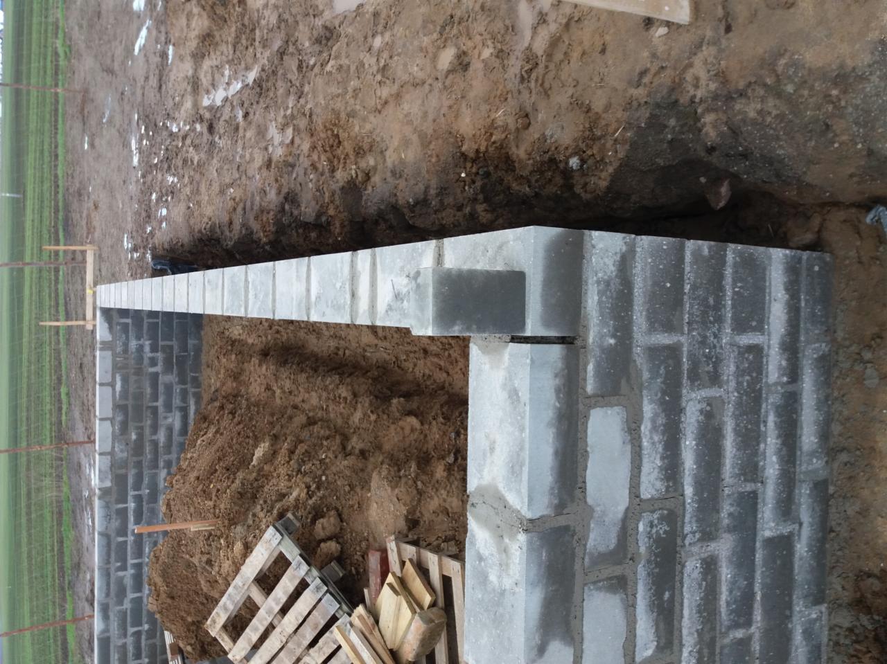 Wykop pod fundamenty z częściowo wzniesioną ścianą z bloczków betonowych, widoczny paleta z drewnem i ziemia.