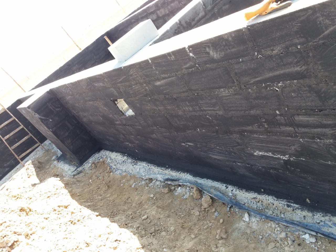 Hydroizolacja pionowa fundamentów z bloczków betonowych, widoczny otwór wentylacyjny i grunt w wykopie.