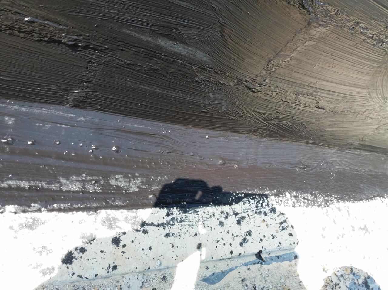 Świeżo nałożona, gruba warstwa czarnej hydroizolacji na dachu, z widocznymi śladami pędzla i odbiciem sylwetki osoby w mokrej powierzchni.