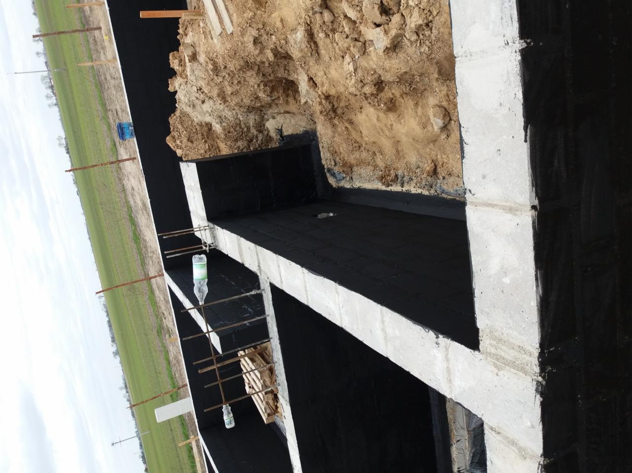 Fundamenty zabezpieczone czarną izolacją bitumiczną w Dobrzejewicach, widoczne zbrojenia, wykop i butelka wody.