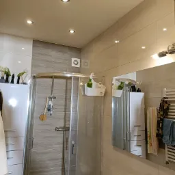 Nowoczesna łazienka z kabiną prysznicową z zaokrąglonymi drzwiami, beżowymi płytkami, białymi szafkami z szufladami, toaletą i akcesoriami łazienkowymi.