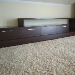 Ciemnobrązowa szafka RTV z czterema szufladami i otwartą półką, z minimalistycznymi srebrnymi uchwytami, na tle kremowej ściany i puszystego, jasnego dywanu, obok stoi czarny głośnik.