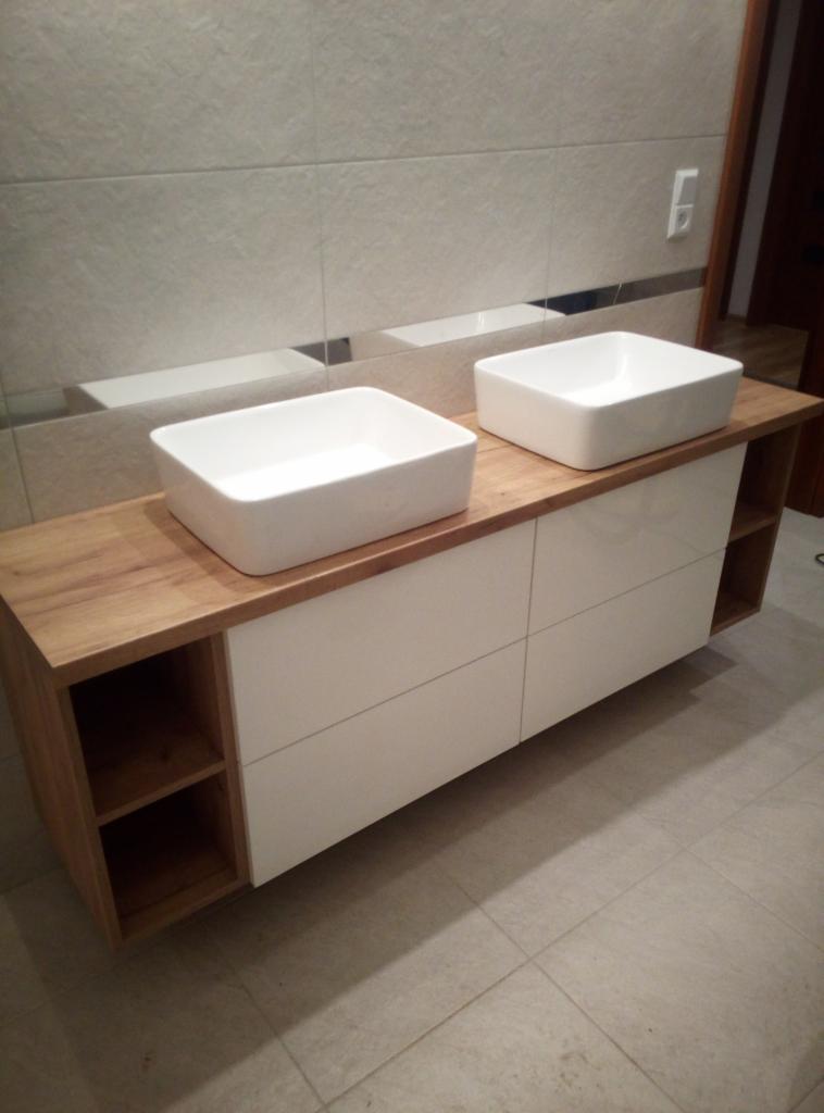 Nowoczesna łazienka z dwiema białymi, prostokątnymi umywalkami nablatowymi, zamontowanymi na szafce z drewnianym blatem i białymi frontami szuflad. Po bokach szafki widoczne otwarte półki. W tle...