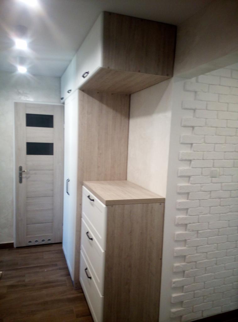 Wnęka z zabudową: szafa z białymi frontami i drewnopodobnymi bokami, obok ściana z białej cegły dekoracyjnej, drzwi w tle.