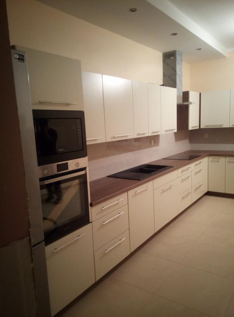 Jasna kuchnia na wymiar z drewnianym blatem, zabudowanym piekarnikiem i mikrofalówką, czarnym zlewozmywakiem i metalowym okapem nad płytą grzewczą.