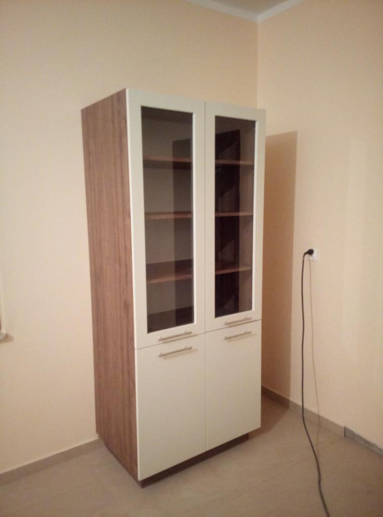 Dwudrzwiowa witryna z drewnianymi bokami i białymi frontami, z przeszkloną górną częścią i półkami wewnątrz, ustawiona w jasnym pomieszczeniu z widocznym gniazdkiem elektrycznym.