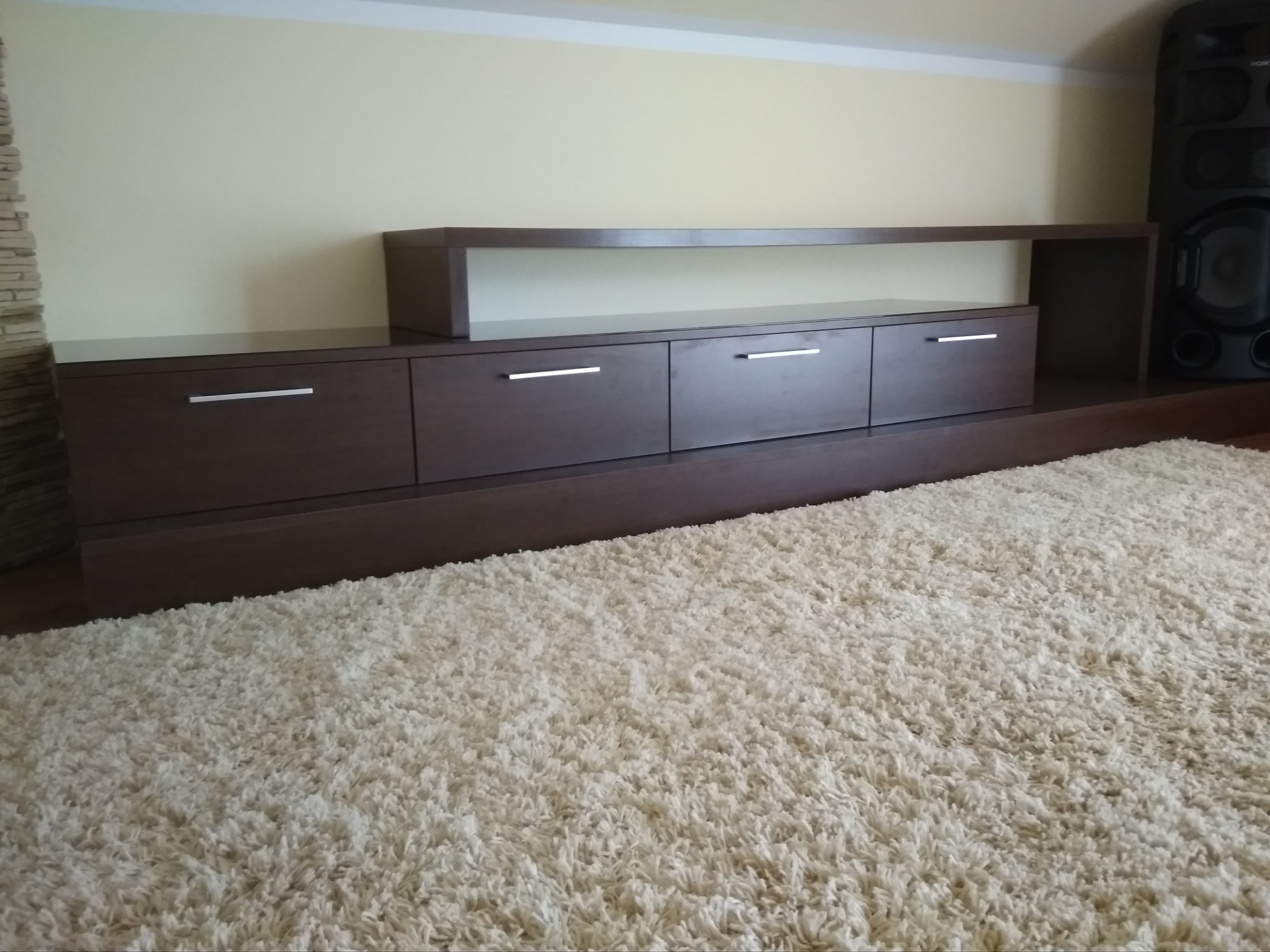 Ciemnobrązowa szafka RTV z czterema szufladami i otwartą półką, z minimalistycznymi srebrnymi uchwytami, na tle kremowej ściany i puszystego, jasnego dywanu, obok stoi czarny głośnik.