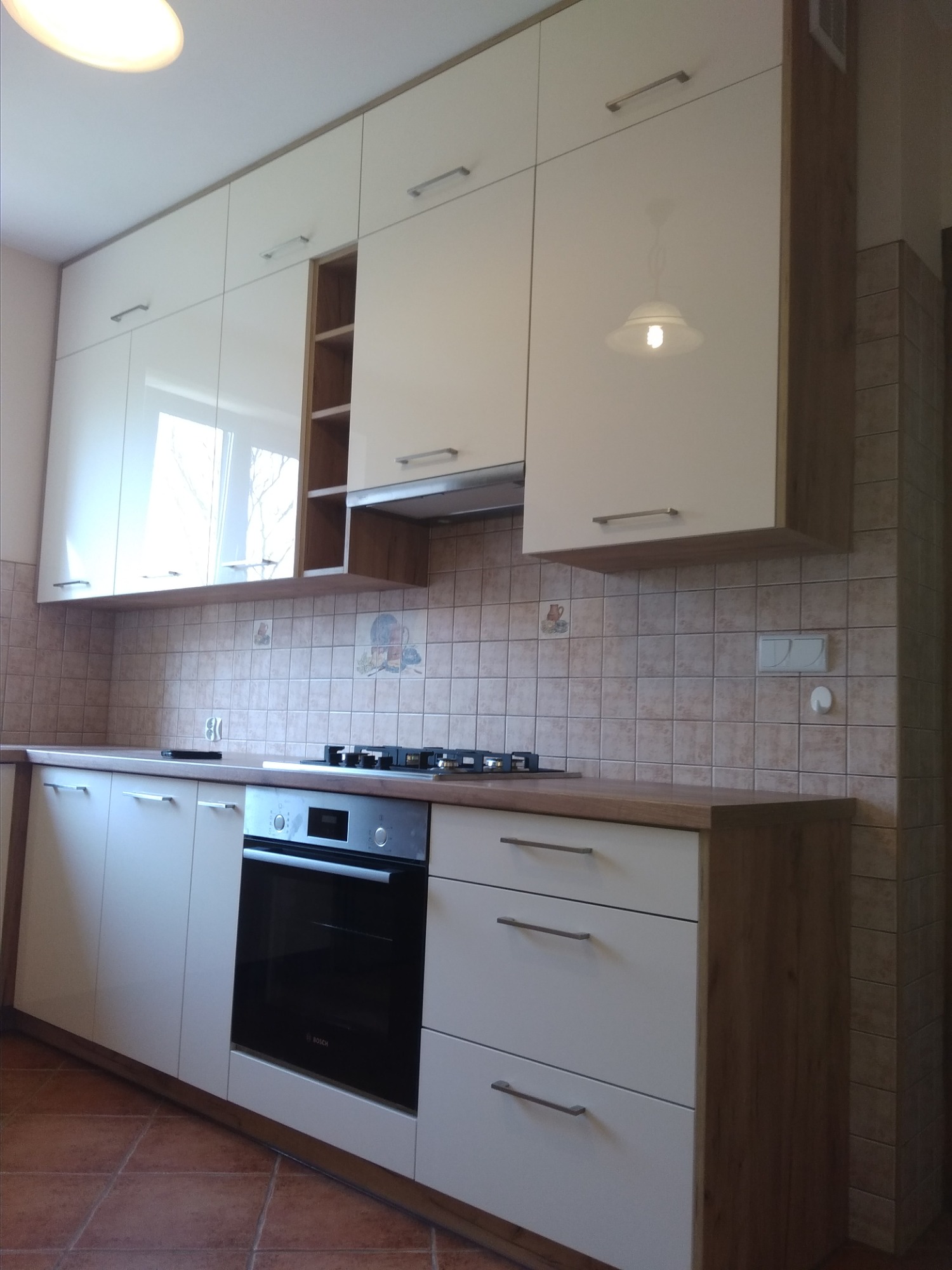 Jasna kuchnia z białymi, lakierowanymi frontami szafek i drewnianymi blatami, widoczna płyta gazowa i piekarnik Bosch, brązowe płytki na ścianie i podłodze.